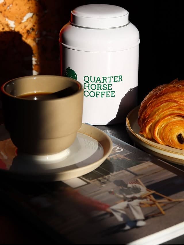 A white Ironroot Coffee tin beside an espresso and a croissant